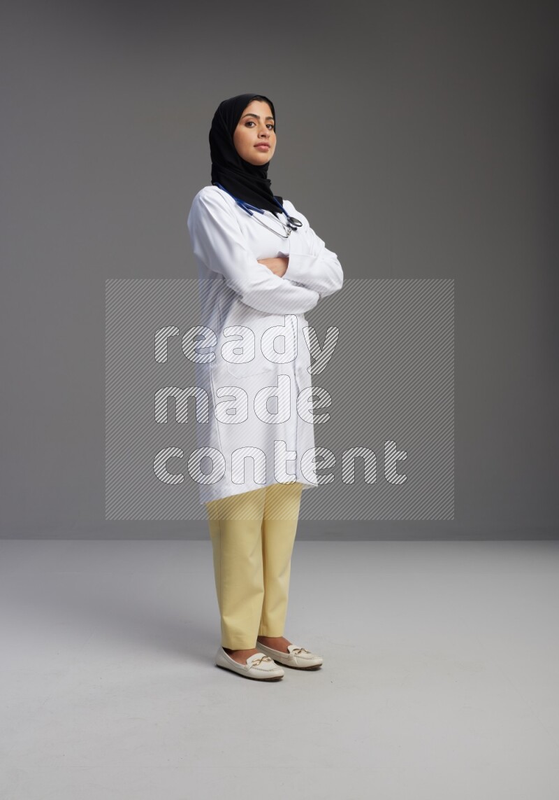 Saudi woman wearing lab coat with stethoscope standing with crossed arms on Gray background