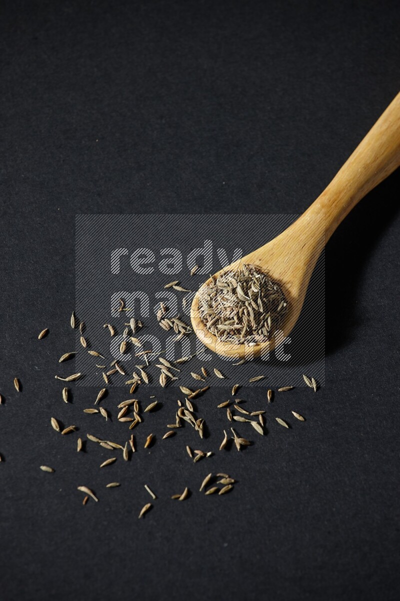 A wooden spoon full of cumin seeds on black flooring