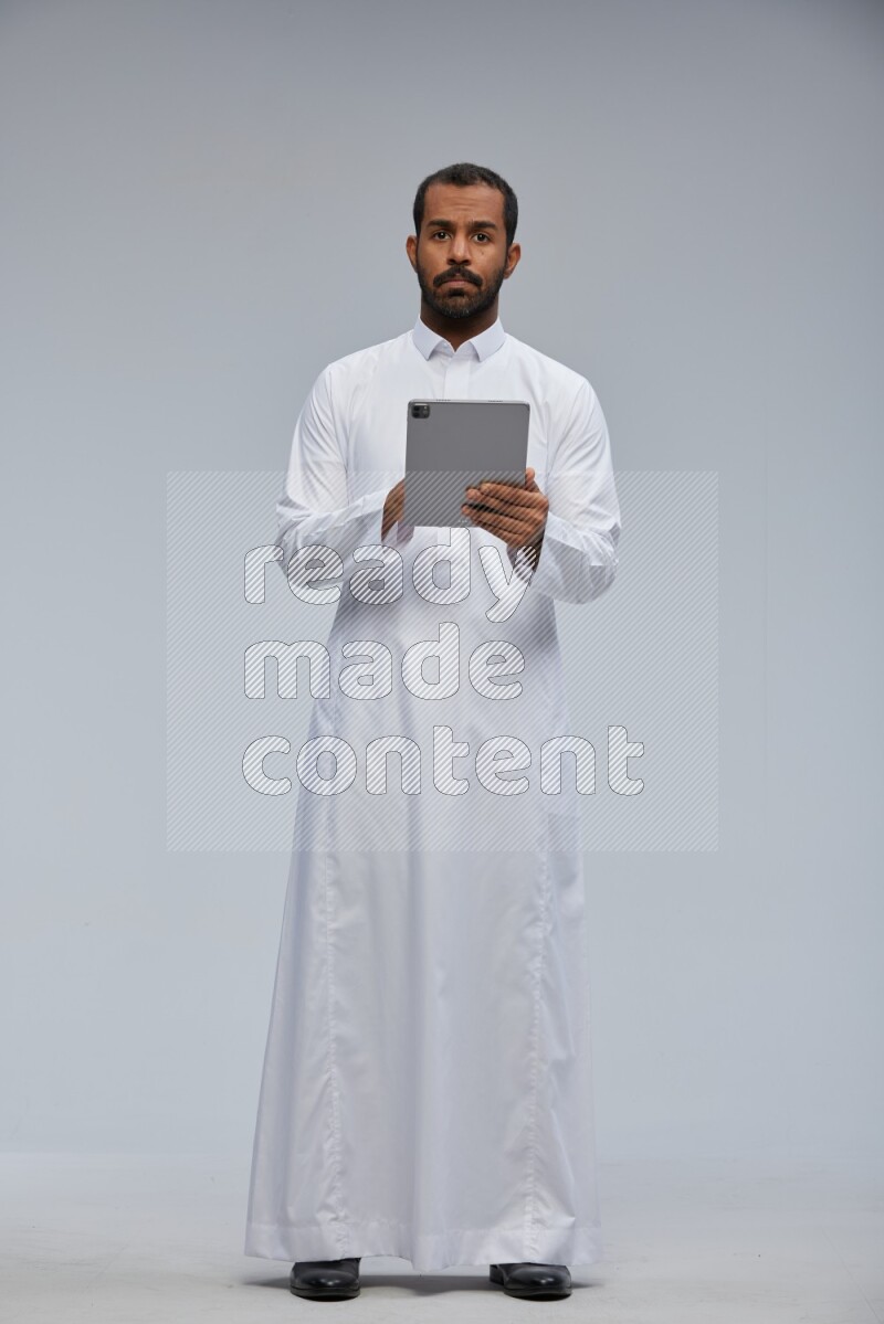 Saudi man wearing Thob standing working on tablet on Gray background