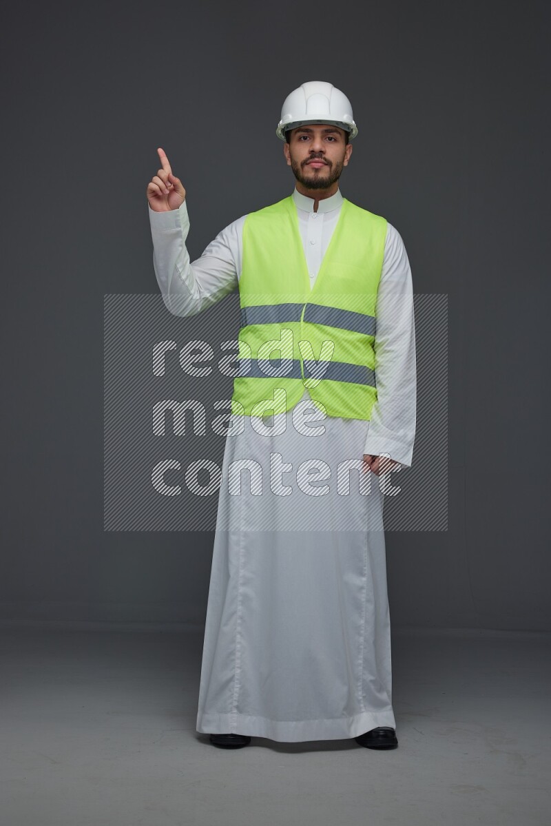 A Saudi man wearing Thobe with a yellow safety vest and white helmet standing and pointing different angles eye level on a gray background
