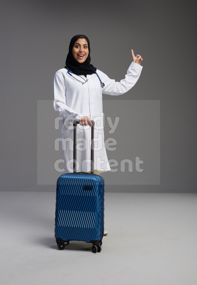 Saudi woman wearing lab coat with stethoscope standing holding Travel bag on Gray background