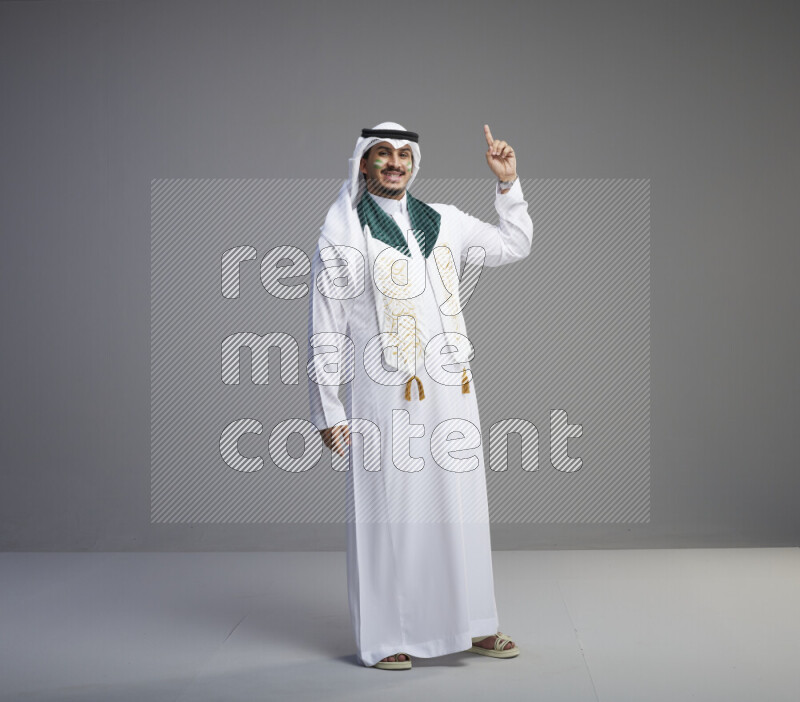 A Saudi man standing wearing thob and white shomag with face painting and Saudi flag scarf on gray background