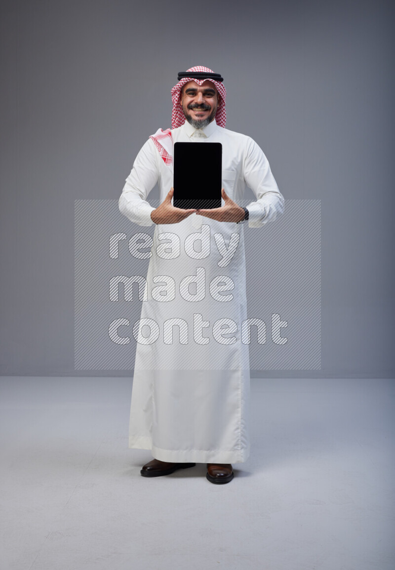 Saudi man Wearing Thob and red Shomag standing showing tablet to camera on Gray background
