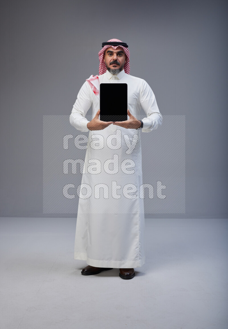 Saudi man Wearing Thob and red Shomag standing showing tablet to camera on Gray background