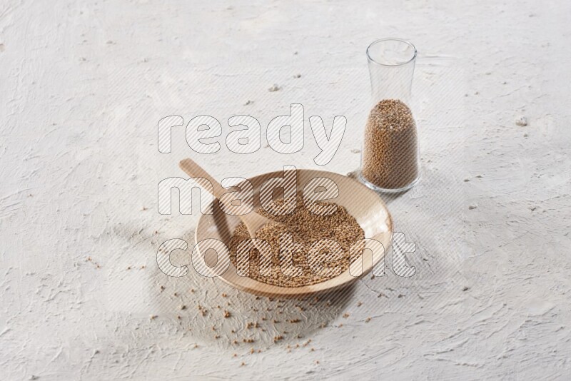 A beige pottery plate full of mustard seeds and a wooden spoon in it with a glass jar filled with the seeds on a textured white flooring