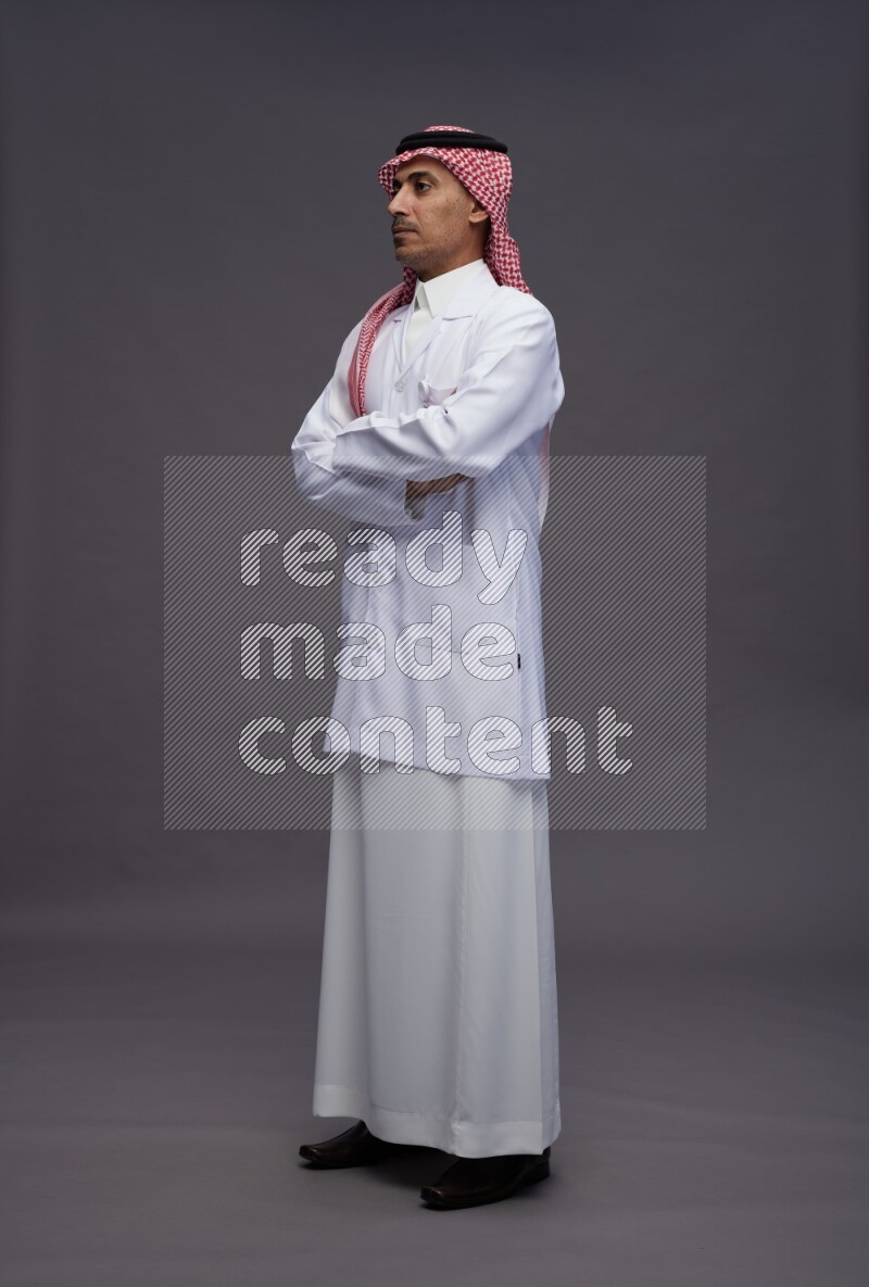 Saudi man wearing thob with lab coat and shomag with pocket employee badge standing with crossed arms on gray background