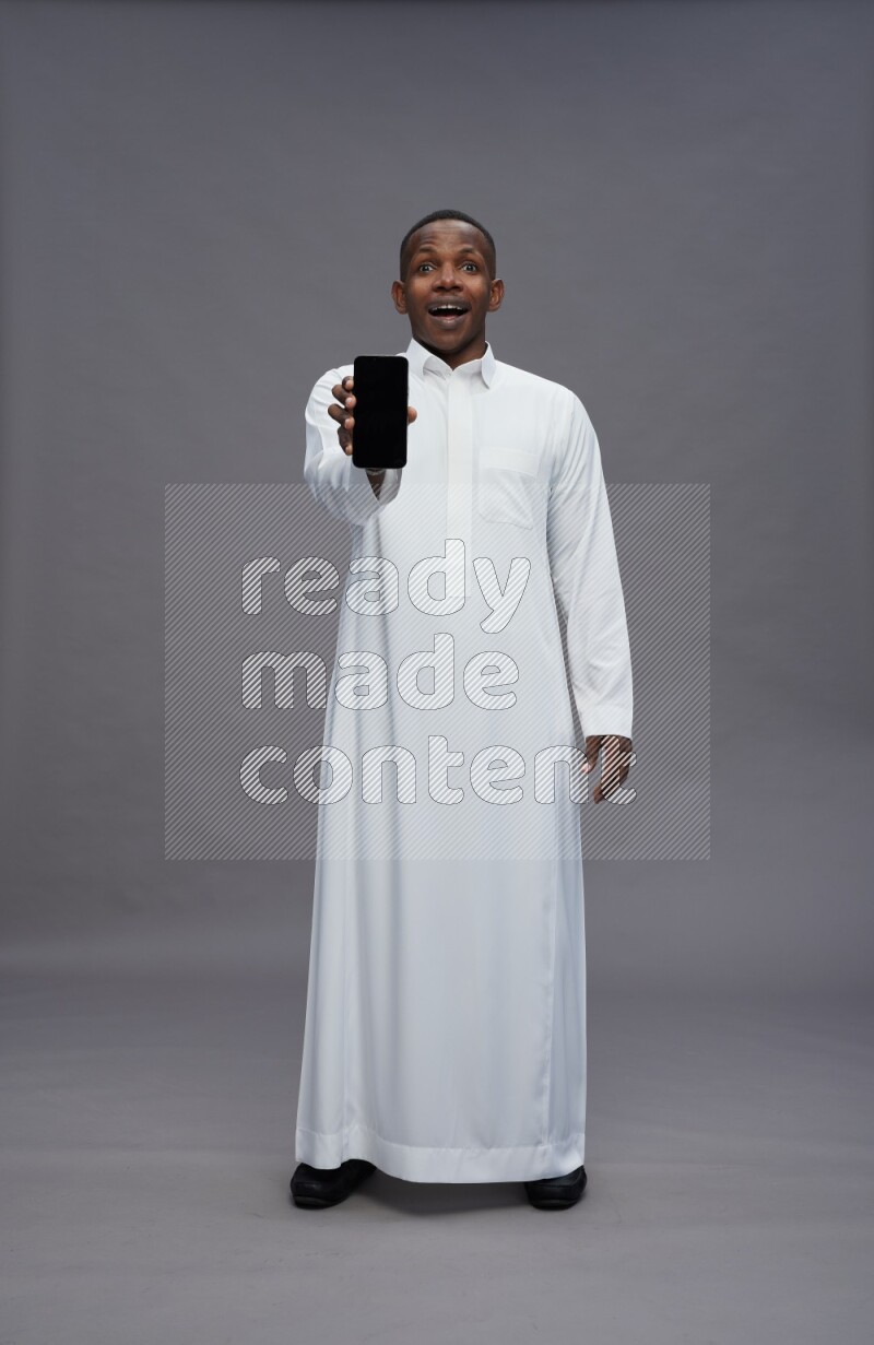 Saudi man wearing thob standing showing phone to camera on gray background