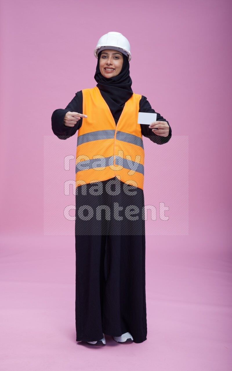 Saudi woman wearing Abaya with engineer vest and helmet standing holding ATM card on pink background