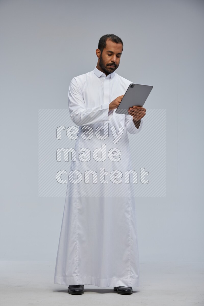Saudi man wearing Thob standing working on tablet on Gray background