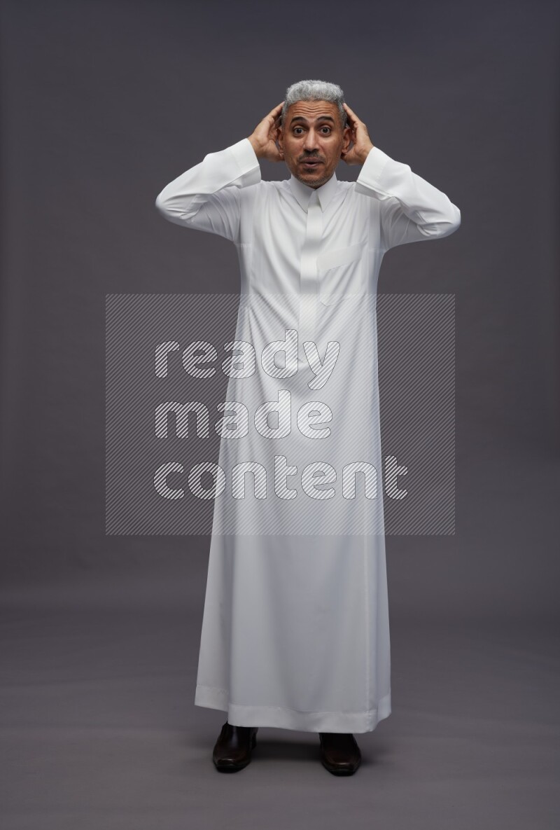 Saudi man wearing thob standing hands behind head on gray background