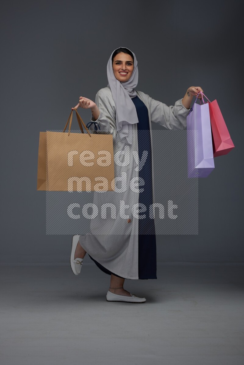 A Saudi woman wearing a light gray Abaya and head scarf standing and holding shopping bags on a grey background