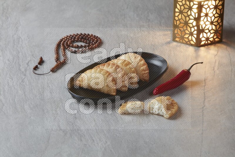 Four fried sambosas in an oval shaped black plate, beside a cut cheese sambosa, a brown misbaha and a golden lantern on a gray background