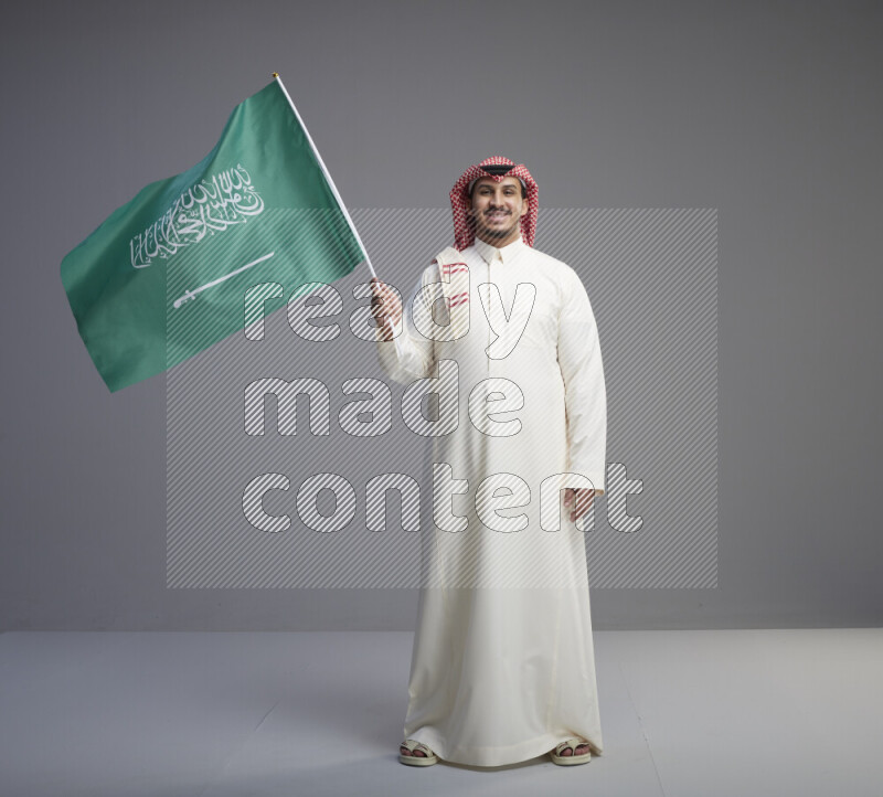 A Saudi man standing wearing thob and red shomag raising big Saudi flag on gray background