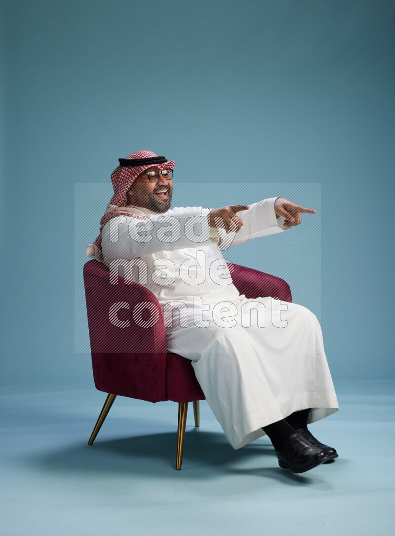 Saudi Man with shimag sitting on chair Interacting with the camera on blue background