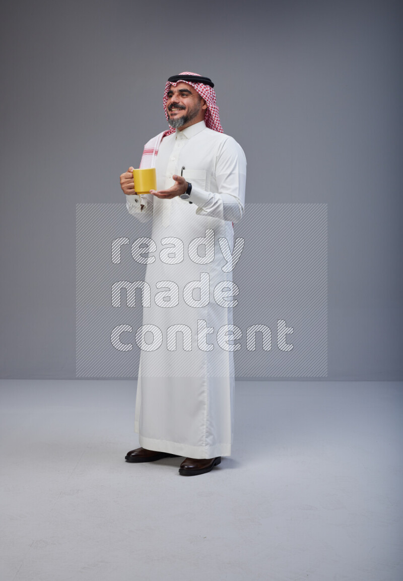Saudi man Wearing Thob and red Shomag standing holding mug on Gray background