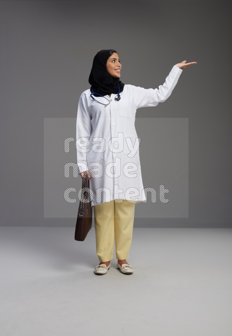 Saudi woman wearing lab coat with stethoscope standing holding bag on Gray background