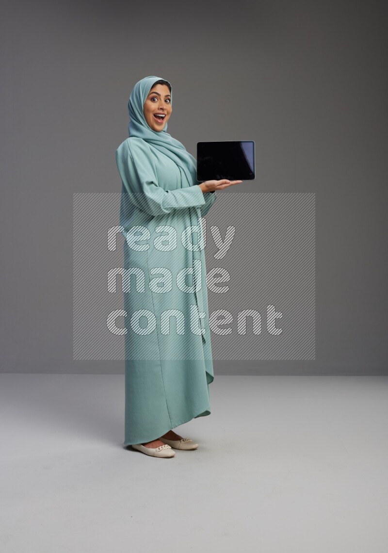 Saudi Woman wearing Abaya standing showing tablet to camera on Gray background