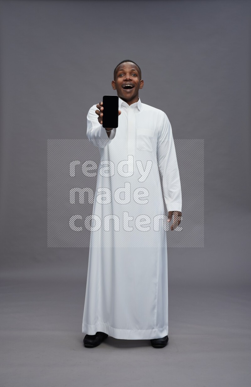 Saudi man wearing thob standing showing phone to camera on gray background