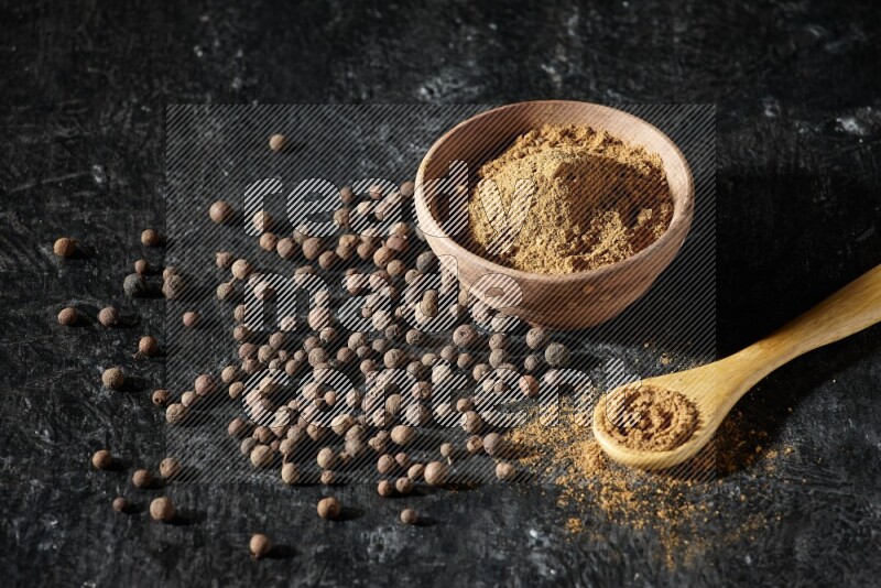 A wooden bowl and spoon full of allspice powder and whole balls spreaded on a black flooring