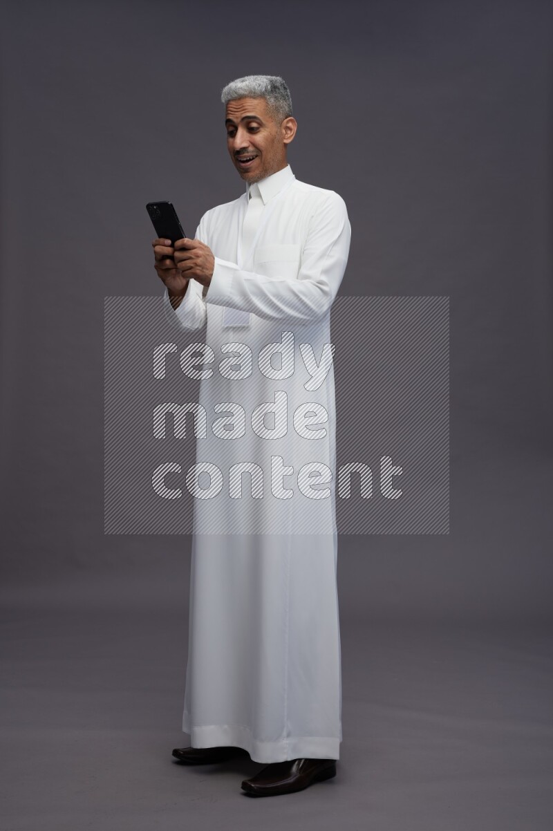 Saudi man wearing thob with neck strap employee badge standing texting on phone on gray background