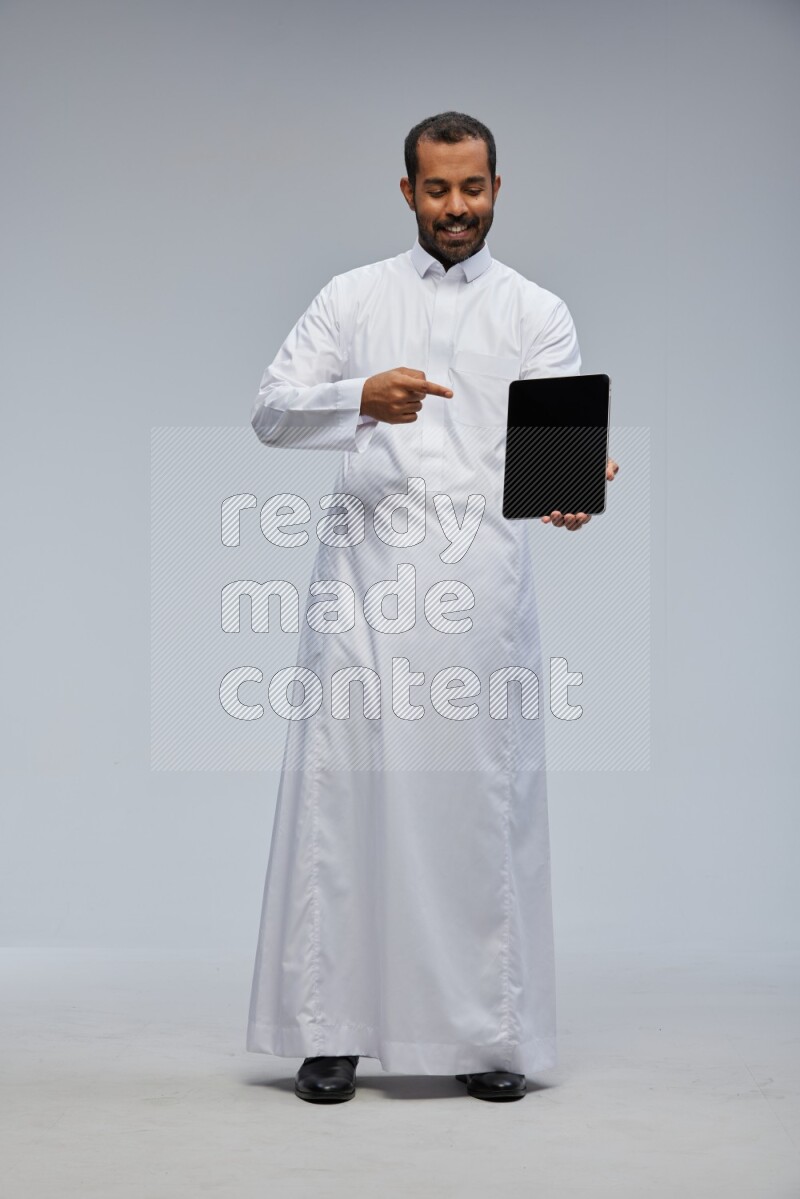 Saudi man wearing Thob standing showing tablet to camera on Gray background