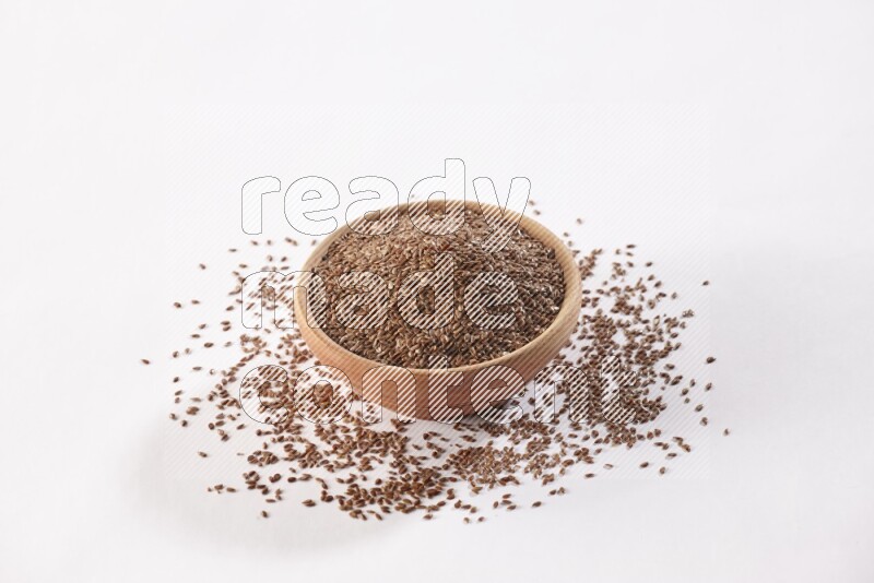 A wooden bowl full of flax seeds surrounded by flax seeds on a white flooring