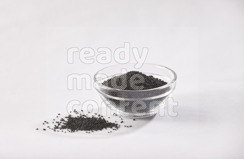 A glass bowl full of black seeds and some more seeds spread next to it on a textured white flooring
