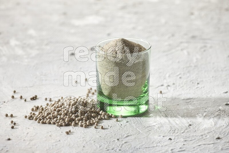 A green glass cup full of white pepper powder with white pepper beads on textured white flooring