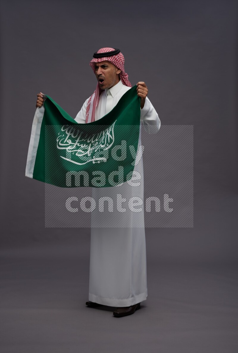 Saudi man wearing thob and shomag standing holding Saudi flag on gray background