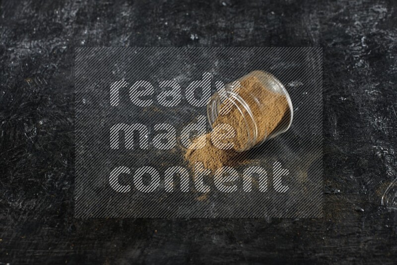 A flipped glass jar full of cumin powder and powder fell out on a textured black flooring