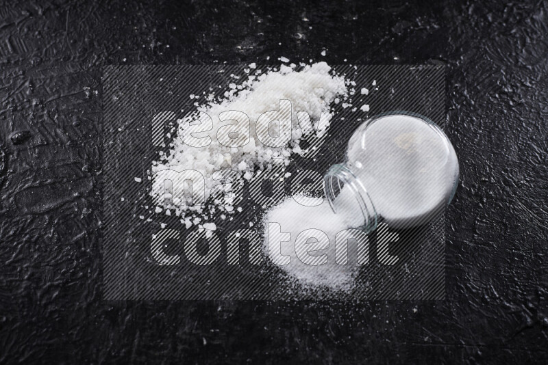 A glass jar full of table salt with some sea salt crystals beside it on a black background