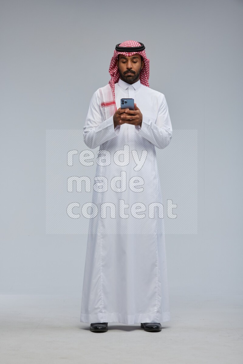Saudi man Wearing Thob and shomag standing texting on phone on Gray background
