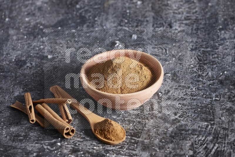 wooden bowl full of cinnamon powder and a wooden spoon full of it with cinnamon sticks on a textured black background