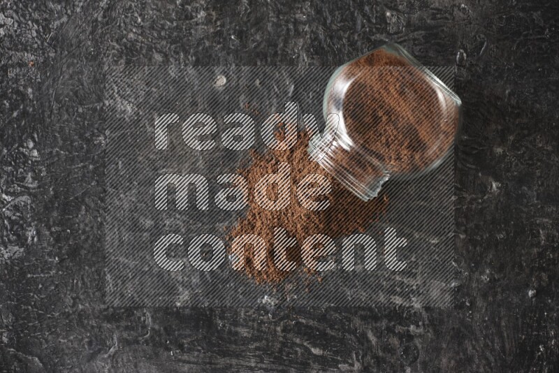 A flipped glass spice jar full of cloves powder and powder came out of it on textured black flooring