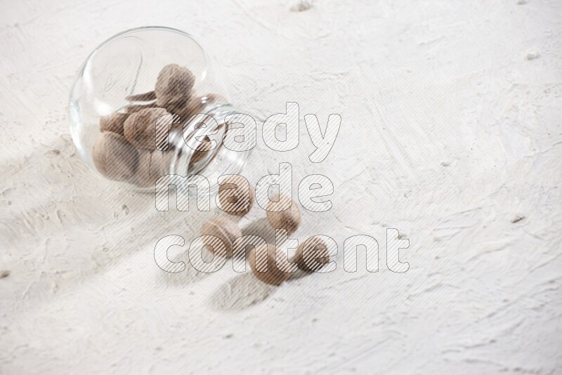A glass spice jar full of whole nutmeg seeds flipped and the seeds spread out on a textured white flooring