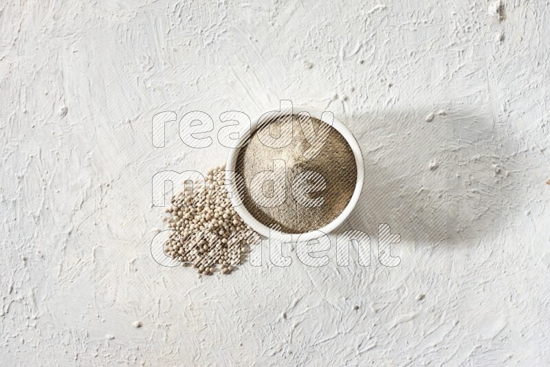 white pottery bowl full of white pepper powder with pepper beads on textured white flooring