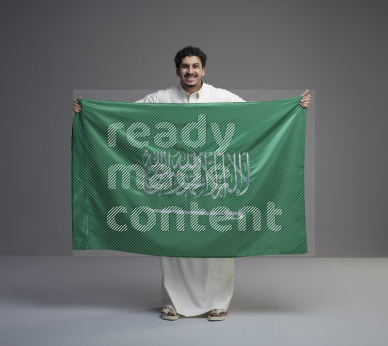 A Saudi man standing wearing thob holding big Saudi flag on gray background