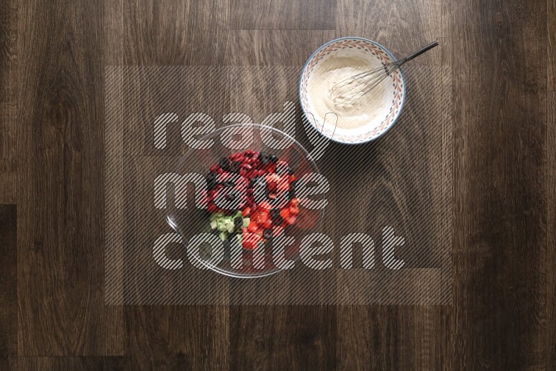 A bowl full of salad (avocado, tomatoes, red beans, olives, bell pepper, corn, lettuce) and bowl of salad dressing on wooden background
