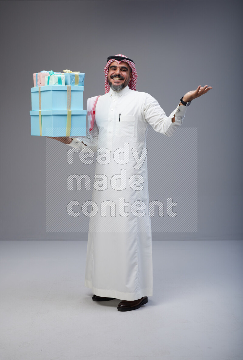 Saudi man Wearing Thob and red Shomag standing holding gift box on Gray background