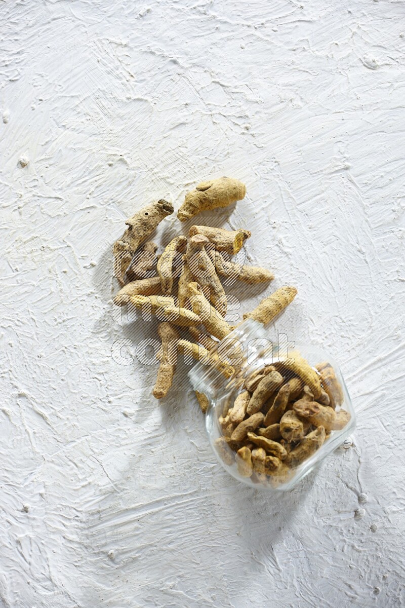 وعاء زجاجي للتوابل مقلوب وممتلئ بأصابع الكركم المجففة وتساقطت خارجه على خلفية بيضاء