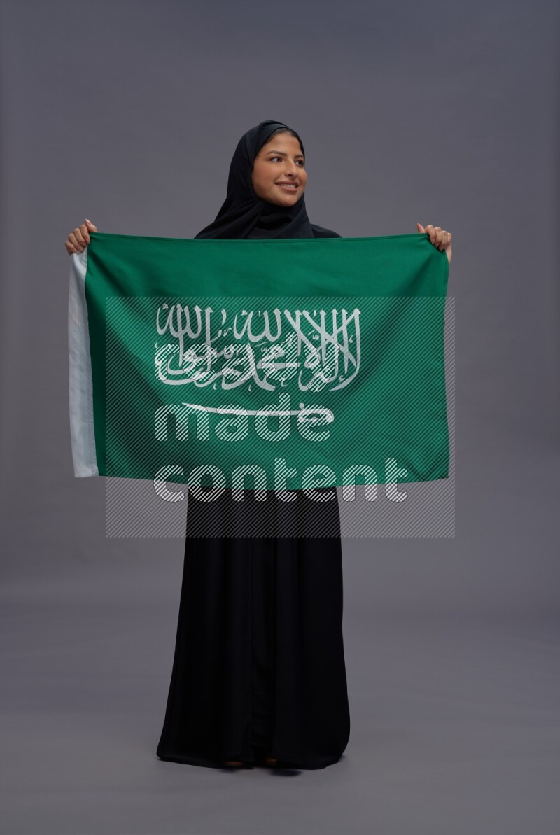 Saudi woman wearing Abaya standing holding Saudi flag on gray background
