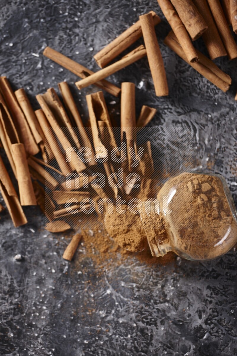Herbal glass jar full cinnamon powder flipped and a metal spoon full of powder surrounded by cinnamon sticks on textured black background in different angles