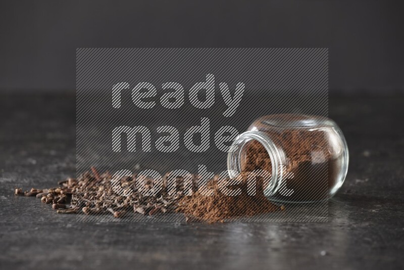 A flipped glass spice jar full of cloves powder and powder came out of it with cloves spread on textured black flooring