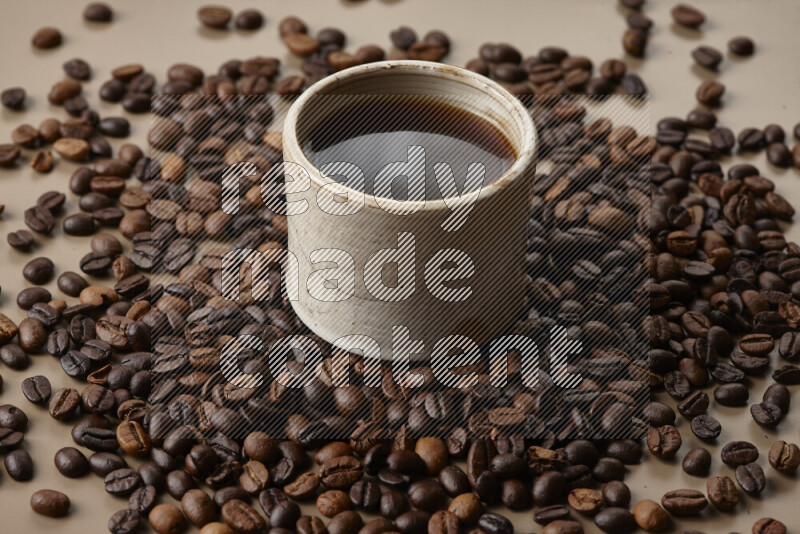 A beige pottery cup of coffee surrounded by roasted coffee beans on beige background