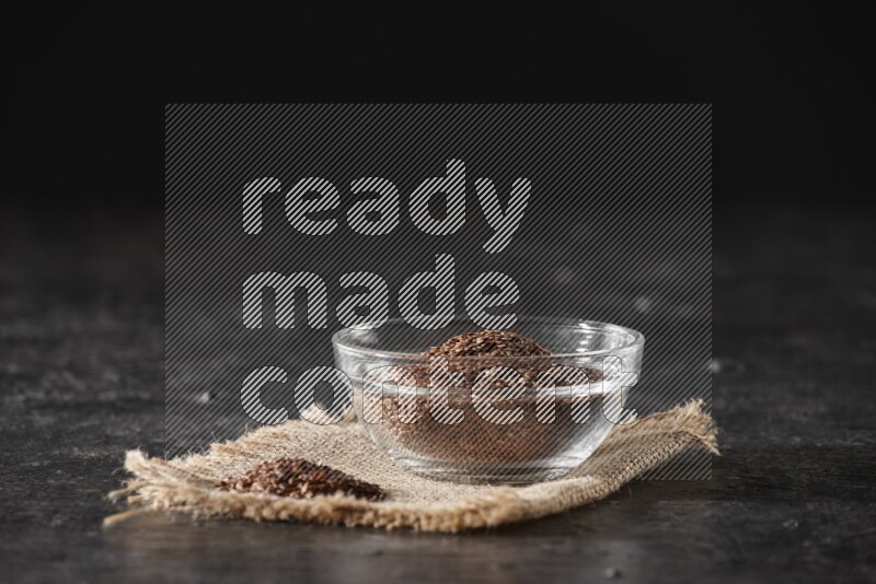 A glass bowl full of flaxseeds with bunch of the seeds on burlap fabric on a textured black flooring