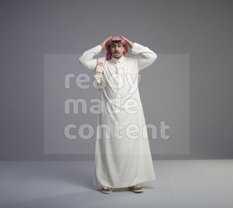A Saudi man standing wearing thob and red shomag interacting with the camera on gray background