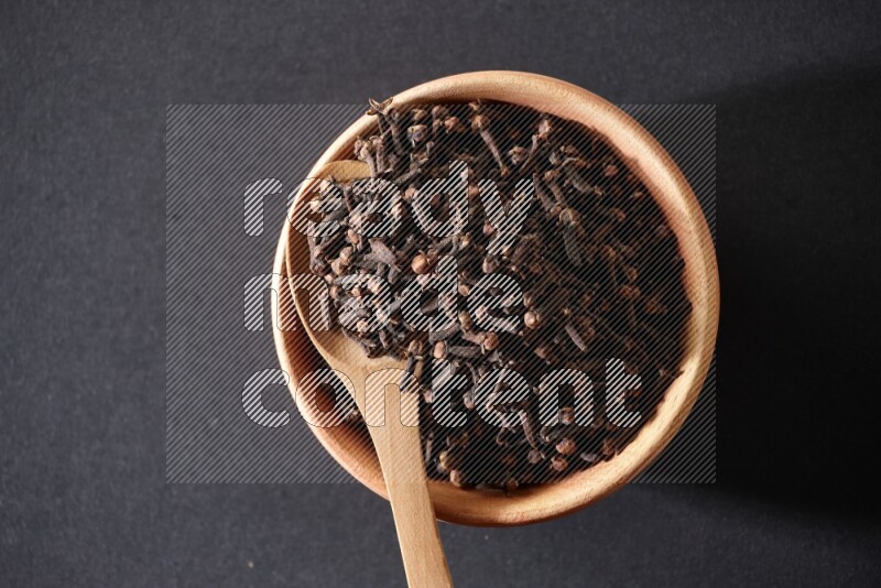 A wooden bowl and a wooden spoon full of cloves on a black flooring
