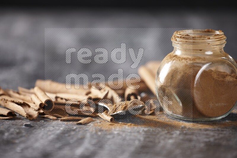 Herbal glass jar and a metal spoon full of cinnamon powder surrounded by cinnamon sticks on textured black background