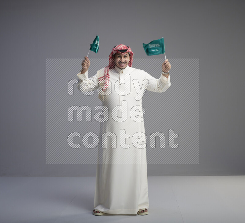 A Saudi man standing wearing thob and red shomag raising small Saudi flag on gray background