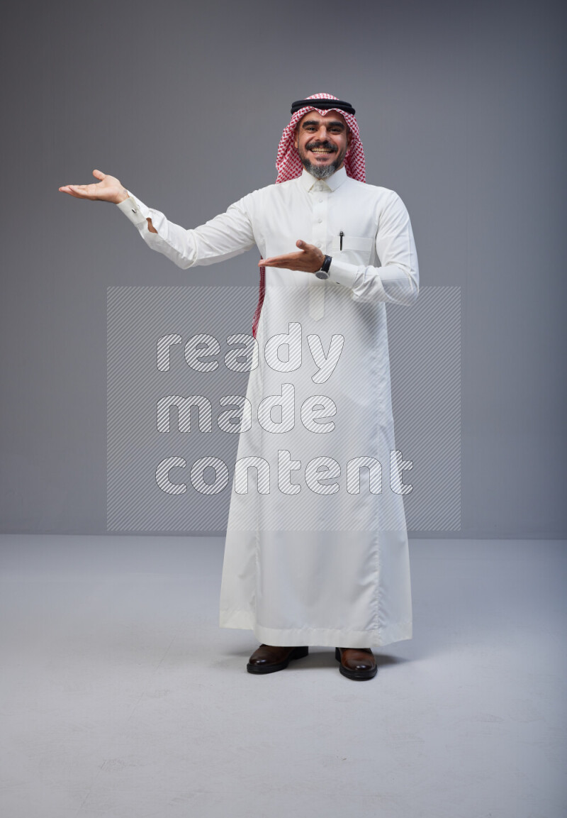 Saudi man Wearing Thob and red white Shomag standing interacting with the camera on Gray background